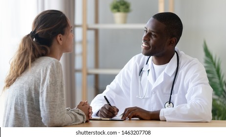 Young Caucasian Woman Talking To Smiling African Black Male Doctor Listening Teen Girl Patient Tell Complaint Making Notes At Medical Checkup Consultation In Clinic, Medicine And Health Care Concept