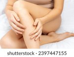 Young Caucasian woman touches her bare hairy legs with her hands. The female sits at home on a white bed. Hair removal, depilation procedures for beauty. Naturalness and body care concept.