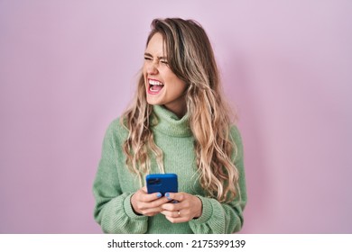 Young Caucasian Woman Using Smartphone Typing Message Angry And Mad Screaming Frustrated And Furious, Shouting With Anger. Rage And Aggressive Concept. 