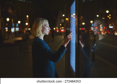 Young caucasian woman using a touch sensitive display while standing on the street with night lights on background, female touching futuristic smart bus station display for self service information