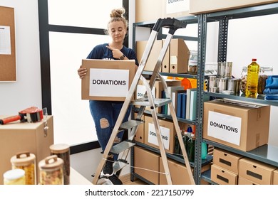 Young Caucasian Woman Volunteer Holding Donations Box Pointing With Hand Finger To The Side Showing Advertisement, Serious And Calm Face 