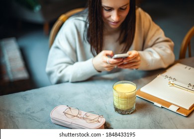 Young caucasian woman in warm sweater is smiling and holding a glass of hot turmeric milk on table of coffee shop. Enjoyment of drinking hot beverage near window on weekend. Copy space
