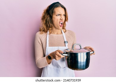Young caucasian woman wearing apron holding cooking pot angry and mad screaming frustrated and furious, shouting with anger. rage and aggressive concept.  - Shutterstock ID 2140731939
