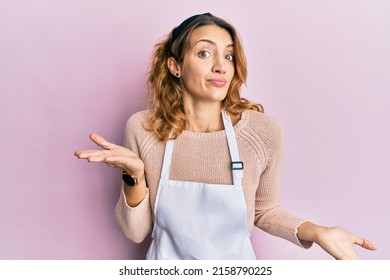 Young Caucasian Woman Wearing Apron Clueless And Confused Expression With Arms And Hands Raised. Doubt Concept. 