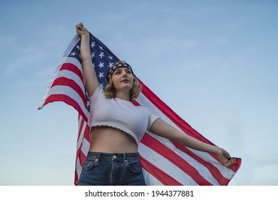 A Young Caucasian Woman Wearing A Bandana And Holding A US Flag Against The Sky