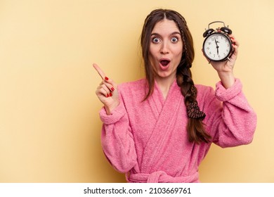 Young caucasian woman wearing a bathrobe holding a alarm clock isolated on yellow background pointing to the side