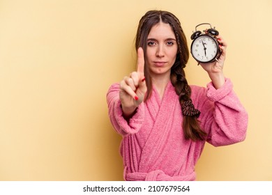 Young caucasian woman wearing a bathrobe holding a alarm clock isolated on yellow background showing number one with finger.