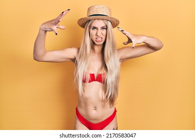 Young caucasian woman wearing bikini and summer hat shouting frustrated with rage, hands trying to strangle, yelling mad 