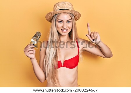 Young caucasian woman wearing bikini eating ice cream smiling with an idea or question pointing finger with happy face, number one 