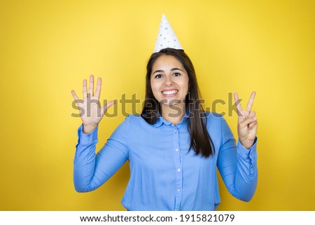 Young caucasian woman wearing a birthday hat over isolated yellow background showing and pointing up with fingers number seven while smiling confident and happy