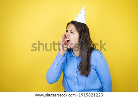 Young caucasian woman wearing a birthday hat over isolated yellow background shouting and screaming loud to side with hand on mouth