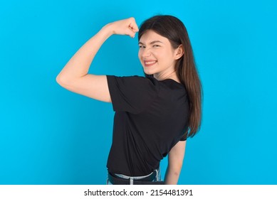 Young caucasian woman wearing black T-shirt over blue background,  showing muscles after workout. Health and strength concept.