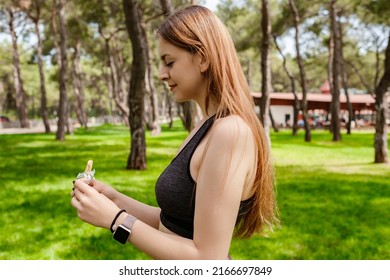 Young Caucasian Woman Wearing Black Sports Bra Standing On City Park, Outdoors Opening Protein Bar Package. Eating Protein Bar, Healthy Food Or Snacks. Green, Sunny Day And Sportive Concepts.