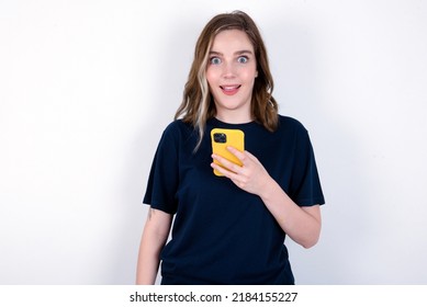 young caucasian woman wearing black T-shirt over white background holds mobile phone in hands and rejoices positive news, uses modern cellular