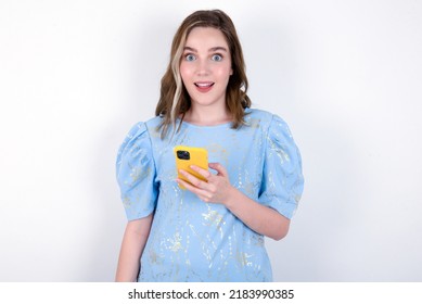 young caucasian woman wearing blue T-shirt over white background holds mobile phone in hands and rejoices positive news, uses modern cellular
