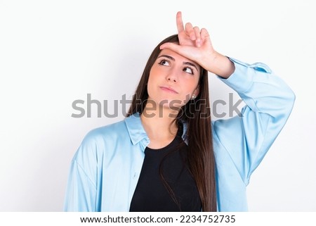 Young caucasian woman wearing blue overshirt over white background making fun of people with fingers on forehead doing loser gesture mocking and insulting.