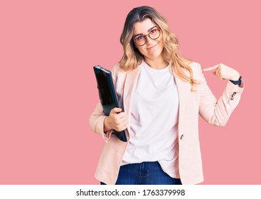 Young caucasian woman wearing business clothes and glasses holding binder pointing finger to one self smiling happy and proud 