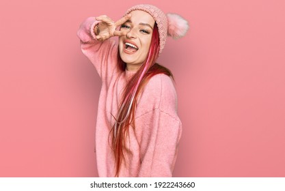 Young caucasian woman wearing casual clothes and wool cap doing peace symbol with fingers over face, smiling cheerful showing victory 