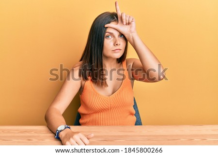 Young caucasian woman wearing casual clothes sitting on the table making fun of people with fingers on forehead doing loser gesture mocking and insulting. 