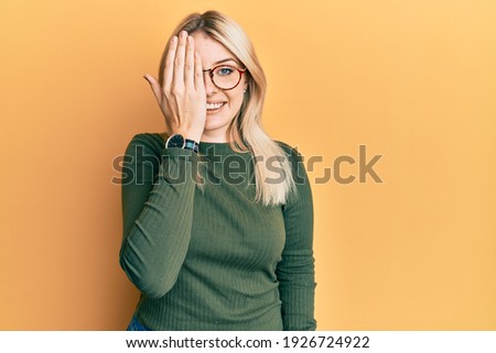 Young caucasian woman wearing casual clothes and glasses covering one eye with hand, confident smile on face and surprise emotion. 