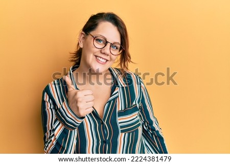 Young caucasian woman wearing casual clothes and glasses doing happy thumbs up gesture with hand. approving expression looking at the camera showing success. 