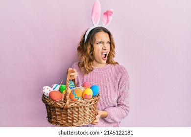 Young caucasian woman wearing cute easter bunny ears holding colored egg angry and mad screaming frustrated and furious, shouting with anger. rage and aggressive concept. 