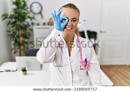 Young caucasian woman wearing doctor uniform and stethoscope at the clinic doing ok gesture with hand smiling, eye looking through fingers with happy face. 