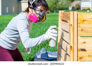 A Young Caucasian Woman Wearing Face Mask Safety Goggles And Gloves Is Spray Staining  A Wooden Fence Outdoors. Concept Image For Diy Project And Health And Safety Measures She Wears Gas Mask.