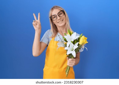 Young caucasian woman wearing florist apron holding flowers smiling looking to the camera showing fingers doing victory sign. number two.  - Powered by Shutterstock