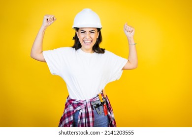 Young caucasian woman wearing hardhat and builder clothes over isolated yellow background very happy and excited making winner gesture with raised arms, smiling and screaming for success.
