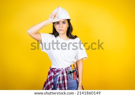 Young caucasian woman wearing hardhat and builder clothes over isolated yellow background making fun of people with fingers on forehead doing loser gesture mocking and insulting.