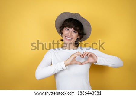 Young caucasian woman wearing hat over isolated yellow background smiling in love showing heart symbol and shape with hands