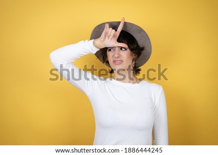 Young caucasian woman wearing hat over isolated yellow background making fun of people with fingers on forehead doing loser gesture mocking and insulting.