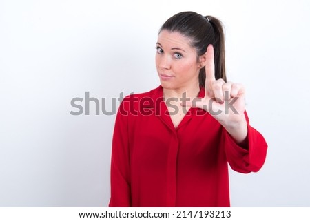 young caucasian woman wearing red shirt over white background making fun of people with fingers on forehead doing loser gesture mocking and insulting.