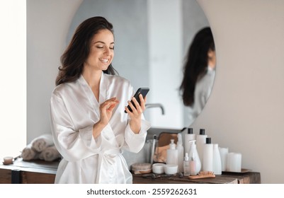 A young Caucasian woman wearing a silk robe smiles as she interacts with her phone, surrounded by skin care products in her stylish bathroom. Its a moment of relaxation and pampering at home. - Powered by Shutterstock