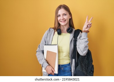 Young caucasian woman wearing student backpack and holding books smiling looking to the camera showing fingers doing victory sign. number two. 