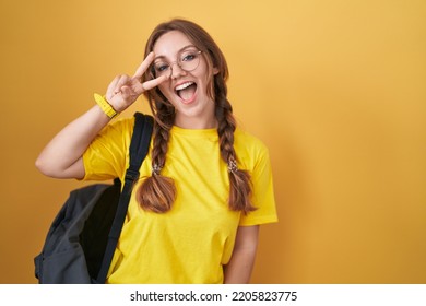 Young caucasian woman wearing student backpack over yellow background doing peace symbol with fingers over face, smiling cheerful showing victory 