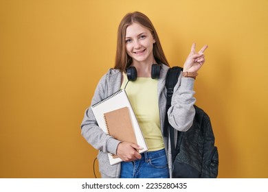 Young caucasian woman wearing student backpack and holding books smiling with happy face winking at the camera doing victory sign with fingers. number two. 