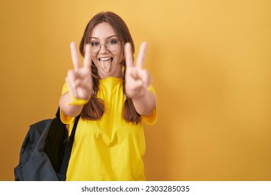 Young caucasian woman wearing student backpack over yellow background smiling with tongue out showing fingers of both hands doing victory sign. number two. 