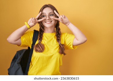 Young caucasian woman wearing student backpack over yellow background doing peace symbol with fingers over face, smiling cheerful showing victory 