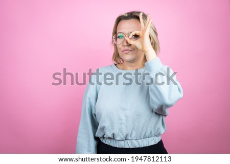 Young caucasian woman wearing sweatshirt over pink background doing ok gesture shocked with smiling face, eye looking through fingers