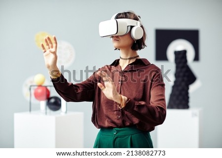 Young Caucasian woman wearing VR headset looking at art objects while visiting modern exhibition using augmented reality technology