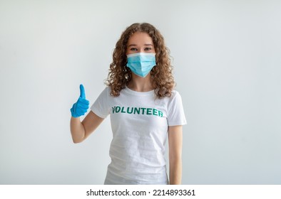 Young Caucasian Woman In White Volunteer T-shirt Showing Thumb Up, Wearing Medical Mask And Gloves, Posing On Light Background, Studio Portrait. Charity Grace Concept