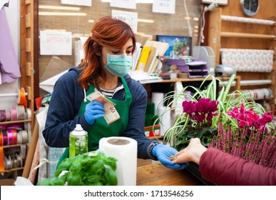 Young caucasian woman at work in a nursery. The florist wears a mask and gloves to avoid the infection of the coronavirus covid-19. The photo shows a customer paying with euro coins and banknotes. - Powered by Shutterstock