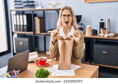 Young caucasian woman working at the office holding paper bag angry screaming frustrated and furious, shouting with anger. rage and aggressive concept.  - Shutterstock ID 2310905997