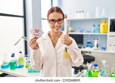 Young Caucasian Woman Working At Scientist Laboratory Holding Toxic Label Smiling Happy And Positive, Thumb Up Doing Excellent And Approval Sign 