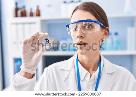 Young caucasian woman working at scientist laboratory holding bitcoin scared and amazed with open mouth for surprise, disbelief face 