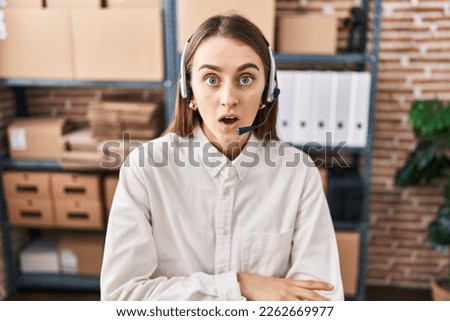 Young caucasian woman working at small business ecommerce wearing headset scared and amazed with open mouth for surprise, disbelief face 