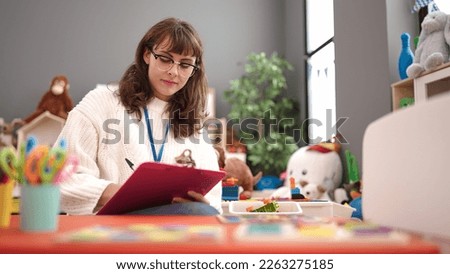 Young caucasian woman working as teacher writing on clipboard at kindergarten
