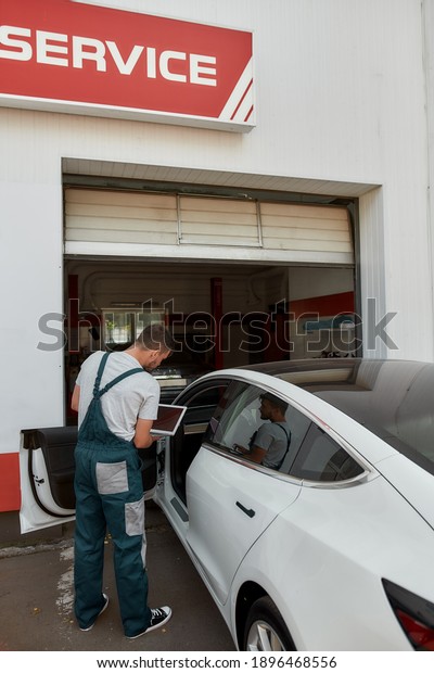 Young caucasian worker doing computer diagnostics\
of electric auto systems on digital laptop on car service station.\
Digital diagnostic of auto, computer applications and repairing\
concept